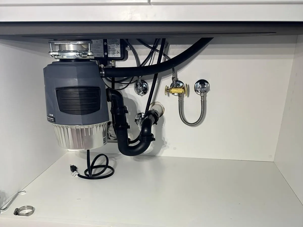 Under a kitchen sink, a new garbage disposal with its black drain trap, two supply valves and the dishwasher line