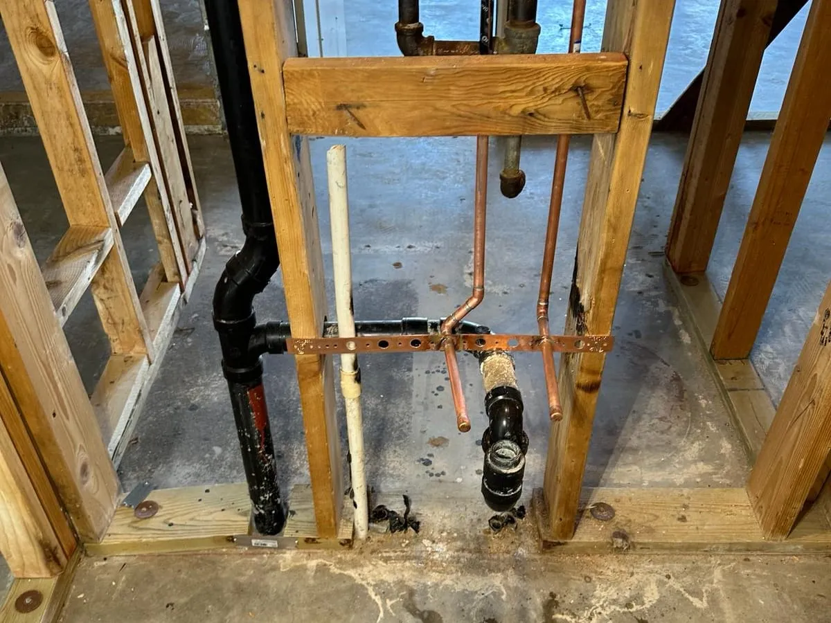 A wall opened down to the studs, showing black drain pipe and new copper water stubs strapped to a bracket above a concrete floor
